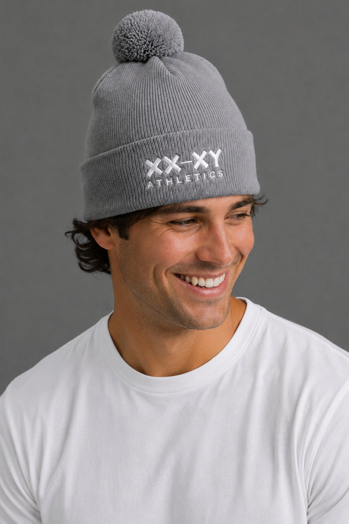A smiling man wearing a gray ribbed knit beanie with a pom-pom and white embroidered 'XX-XY ATHLETICS' text on the front, against a plain gray background. || Heather Gray