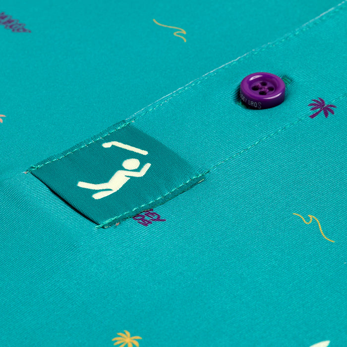 Close-up of the Beach - Polo shirt in teal fabric with an athletic fit, featuring purple palm tree and yellow squiggle prints, a green tag with a white stick figure swimming, and a button detail.