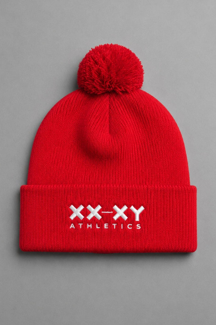 A bright red knit pom-pom beanie with a folded cuff featuring the white embroidered text 'XX-XY ATHLETICS', shown against a solid gray background. || Red