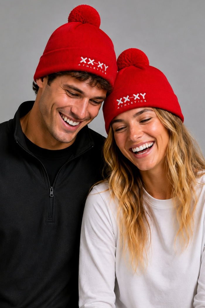 A man and a woman wearing matching red knit pom-pom beanies with white 'XX-XY FANTASY' text on the front, posed against a plain gray studio background. || red