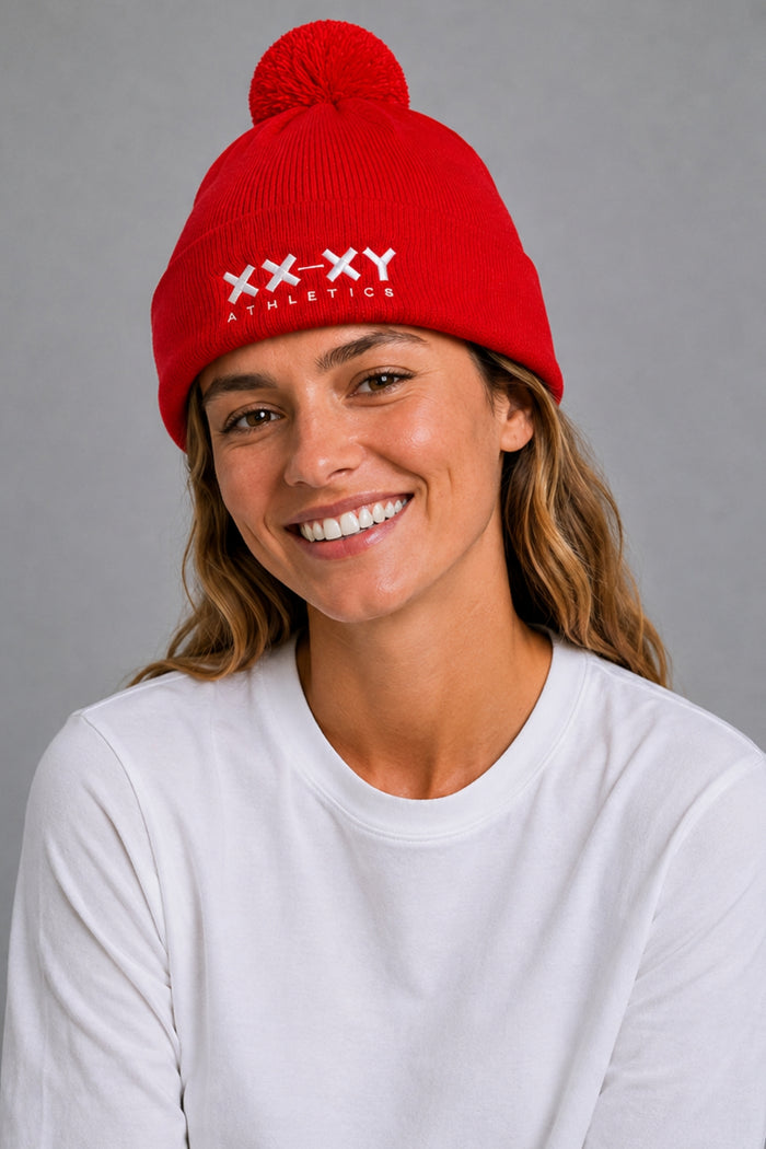 A smiling woman wearing a bright red knit beanie with a pom-pom and white embroidered 'XX-XY ATHLETICS' text on the front, paired with a white long-sleeve shirt against a plain gray studio background. || Red