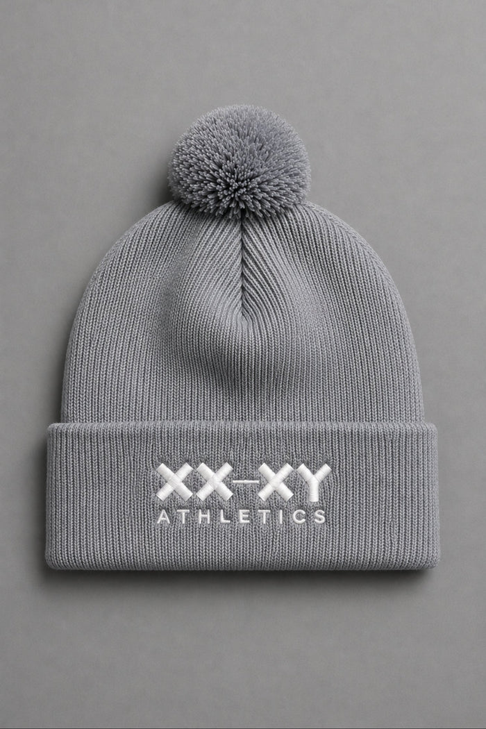 A light gray ribbed knit beanie with a matching pom-pom on top and a folded cuff featuring the white embroidered text 'XX-XY ATHLETICS', shown against a solid gray background. || Heather Gray