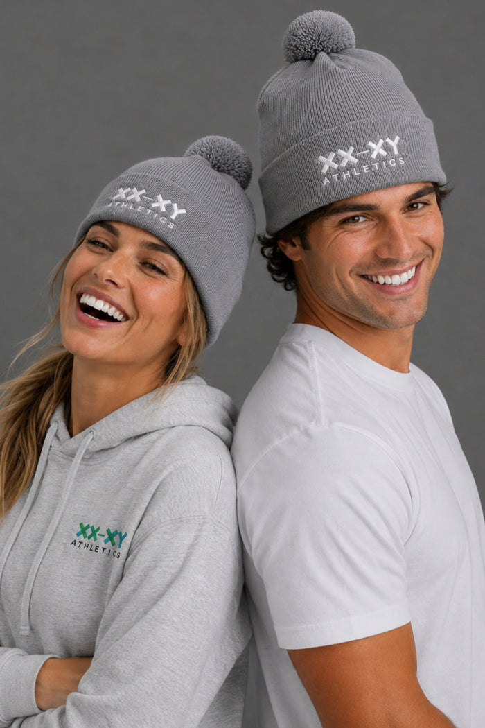 Male and female models wearing gray XX-XY Athletics pom beanies, a gray logo hoodie, and a white T-shirt, posing back-to-back against a gray studio background. || Heather Gray