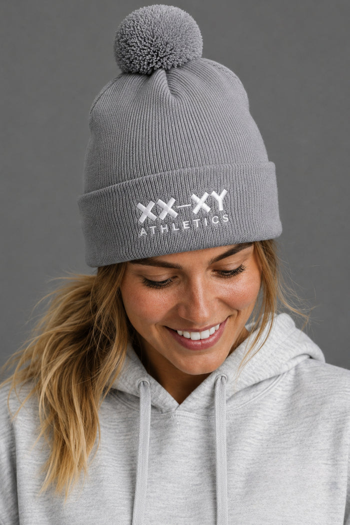 A woman wearing a gray ribbed knit pom-pom beanie with 'XX-XY ATHLETICS' embroidered in white on the front cuff, paired with a light gray hoodie against a dark gray background. || Heather Gray