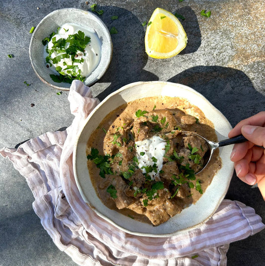 Creamy sauce with chunks of slow cooked grass fed beef, encased in a handmade ceramic white bowl, sprinkled with parsley and a dollop of yoghurt. Placed on a flat grey service, a slice of lemon beside the bowl and a small ramekin beside it holding extra yoghurt.