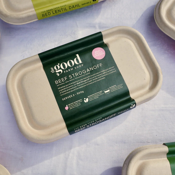 Packaging containing beef stroganoff. A container with soft earthy tones and a dark green label with the good farm shop branding, labelled stroganoff. On a white tablecloth.