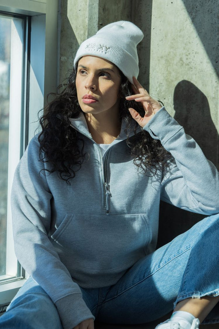 Woman wearing a light blue hoodie and white beanie sitting against a concrete wall. || white