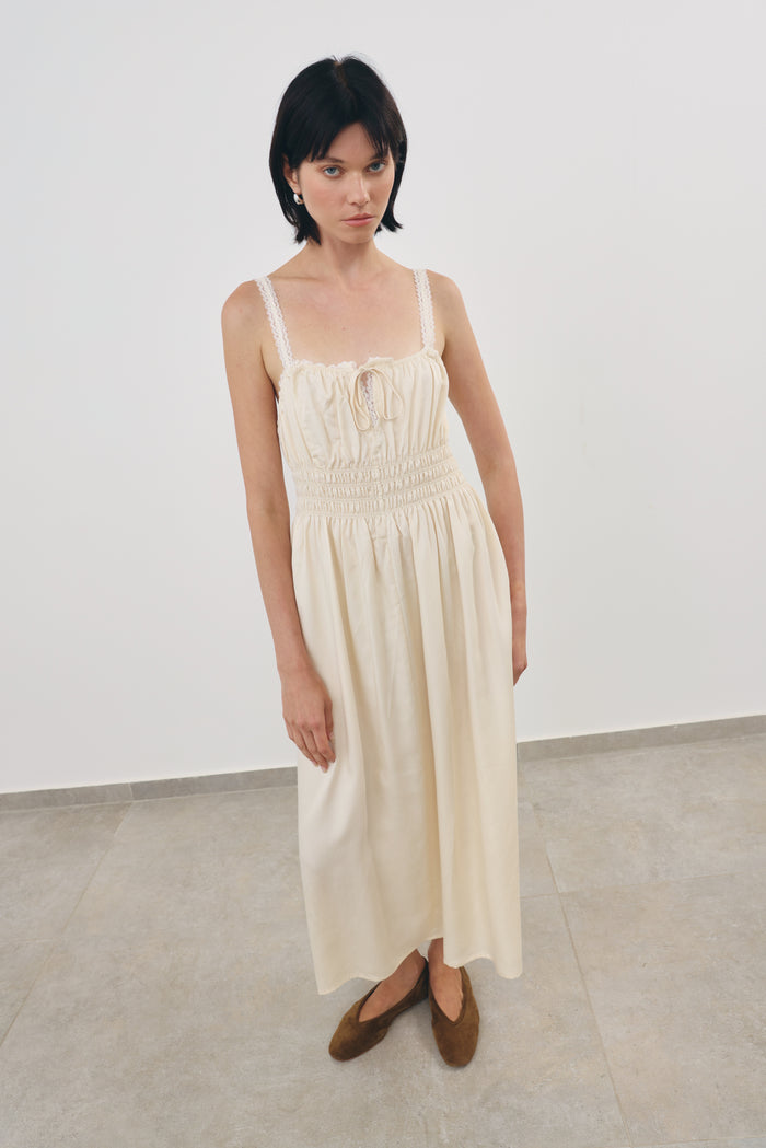A model wearing a cream-colored viscosa midi dress with a smocked bodice and thin lace shoulder straps, paired with khaki flat shoes, standing in a minimalist studio setting.