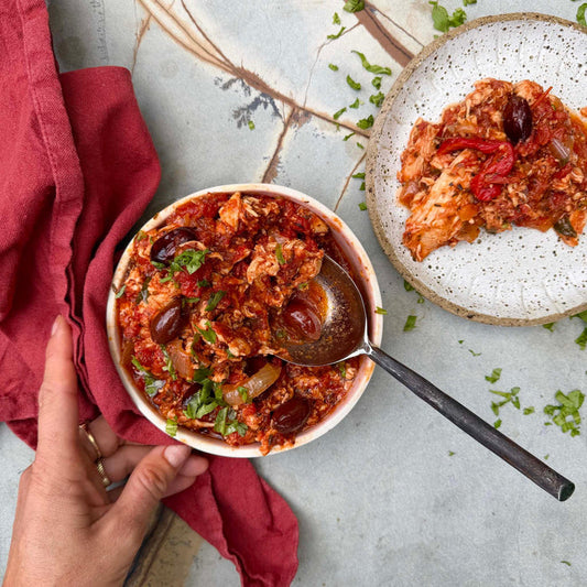 Bowl of hearty chicken and tomato stew with olives with a spoon, placed on a textured surface with a red cloth.