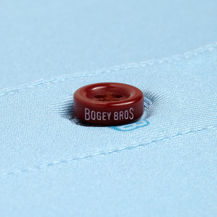 A close-up of a maroon "BOGEY BROS" button sewn onto light blue fabric—Port - Polo, crafted for an athletic fit and designed with moisture-wicking comfort.