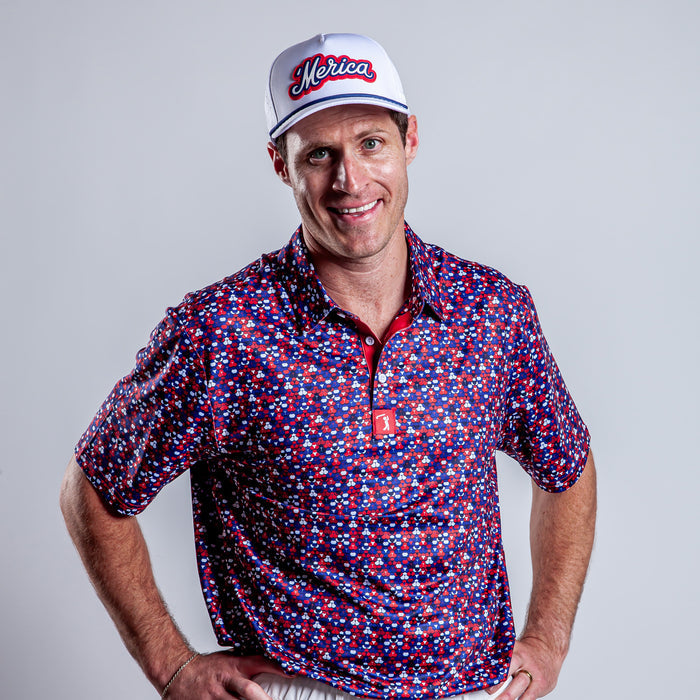 A man, smiling with hands on hips, wears the Whale Tail - Freedom Cheeks - Polo—an athletic fit, short-sleeved button-up with a red, white, and blue star pattern—white "Merica" cap, white pants. Plain light gray background.