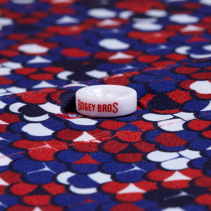 A white golf ball marker with red "BOGEY BROS" text sits centered and in focus on the Whale Tail - Freedom Cheeks - Polo, which features a vibrant pattern of red, blue, and white scalloped shapes.